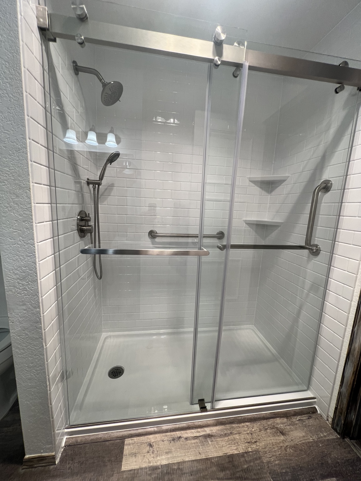 Finished lower-entry shower with white subway-pattern walls, sliding glass doors, grab bars, two showerheads, and matching corner shelves