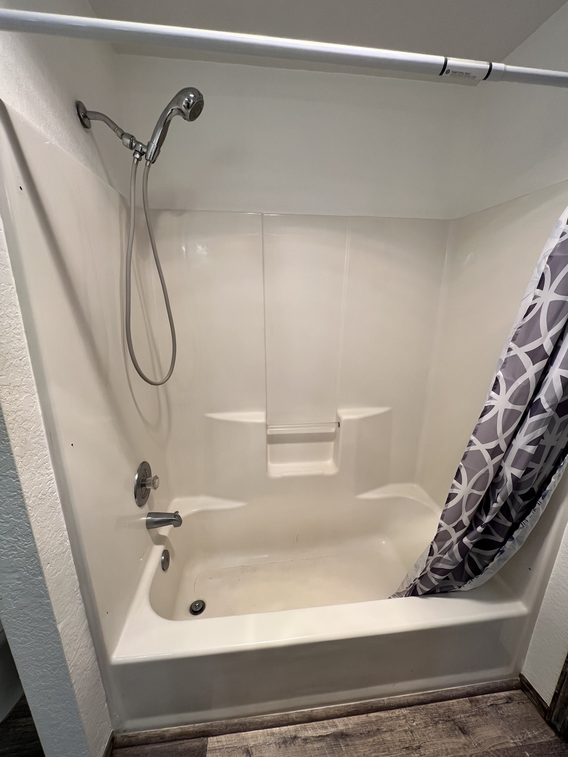 Original bathtub with a cream-colored surround, handheld showerhead, and patterned shower curtain
